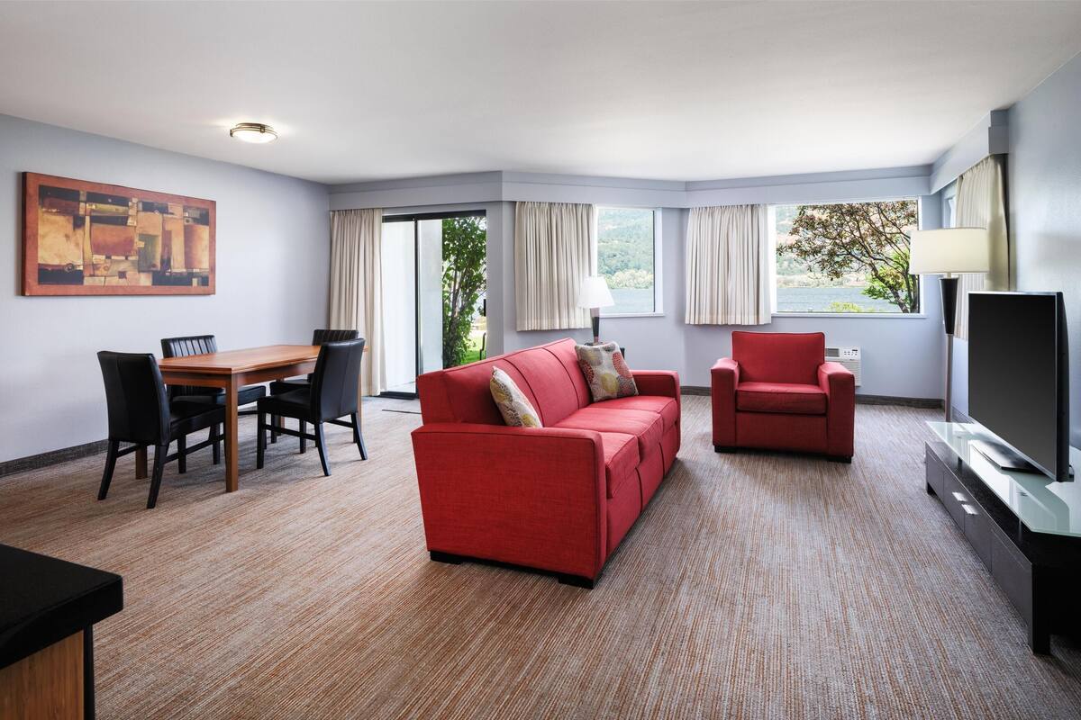 A hotel room with a red sofa, a red armchair, a TV, a dining table with chairs, and a large window with curtains.