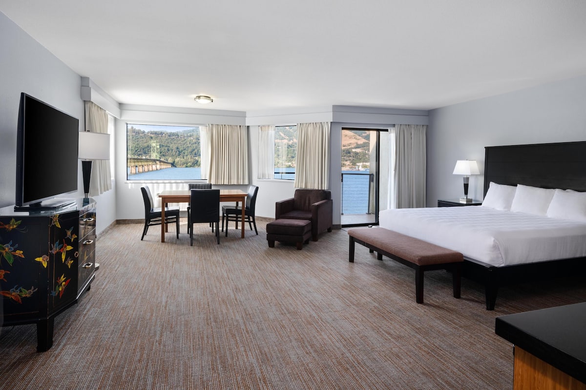 A hotel room with a large bed, a desk with chairs, a television, and a view of a bridge and hills.