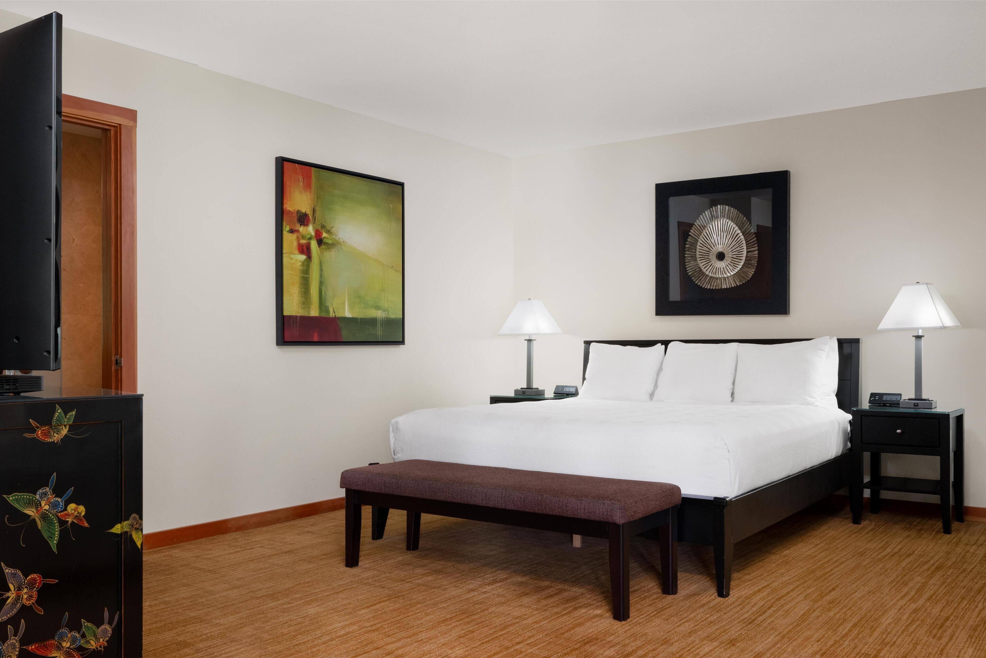 Habitación de hotel con una cama grande, dos mesitas de noche con lámparas, un banco, un televisor en un mueble y dos obras de arte colgadas en la pared.