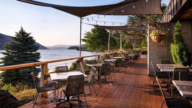 Une terrasse extérieure avec des tables et des chaises, offrant une vue sur un plan d’eau et des collines.