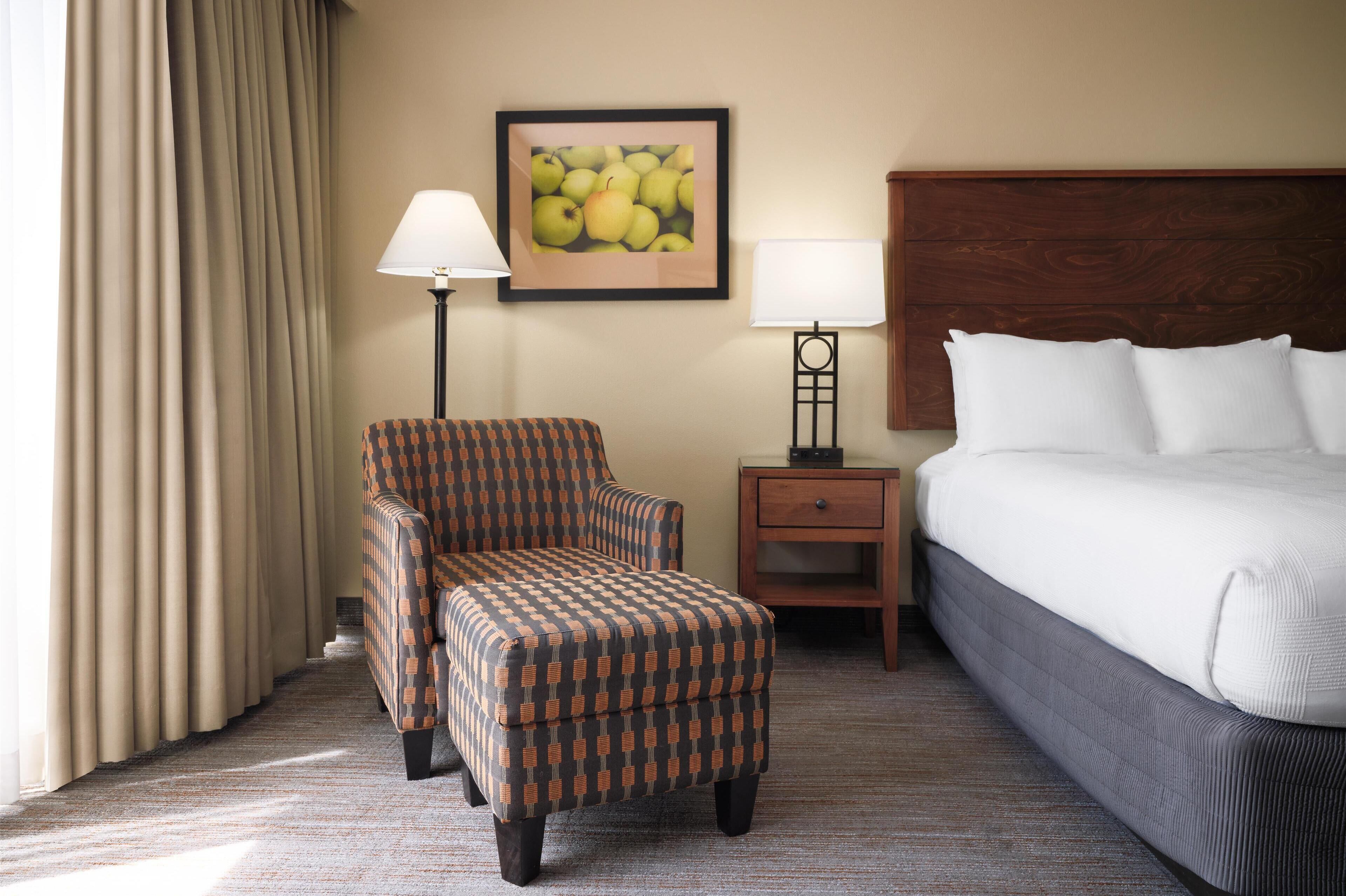 Une chambre d’hôtel avec un lit, une chaise, une table de chevet, une lampe et un tableau encadré au mur.