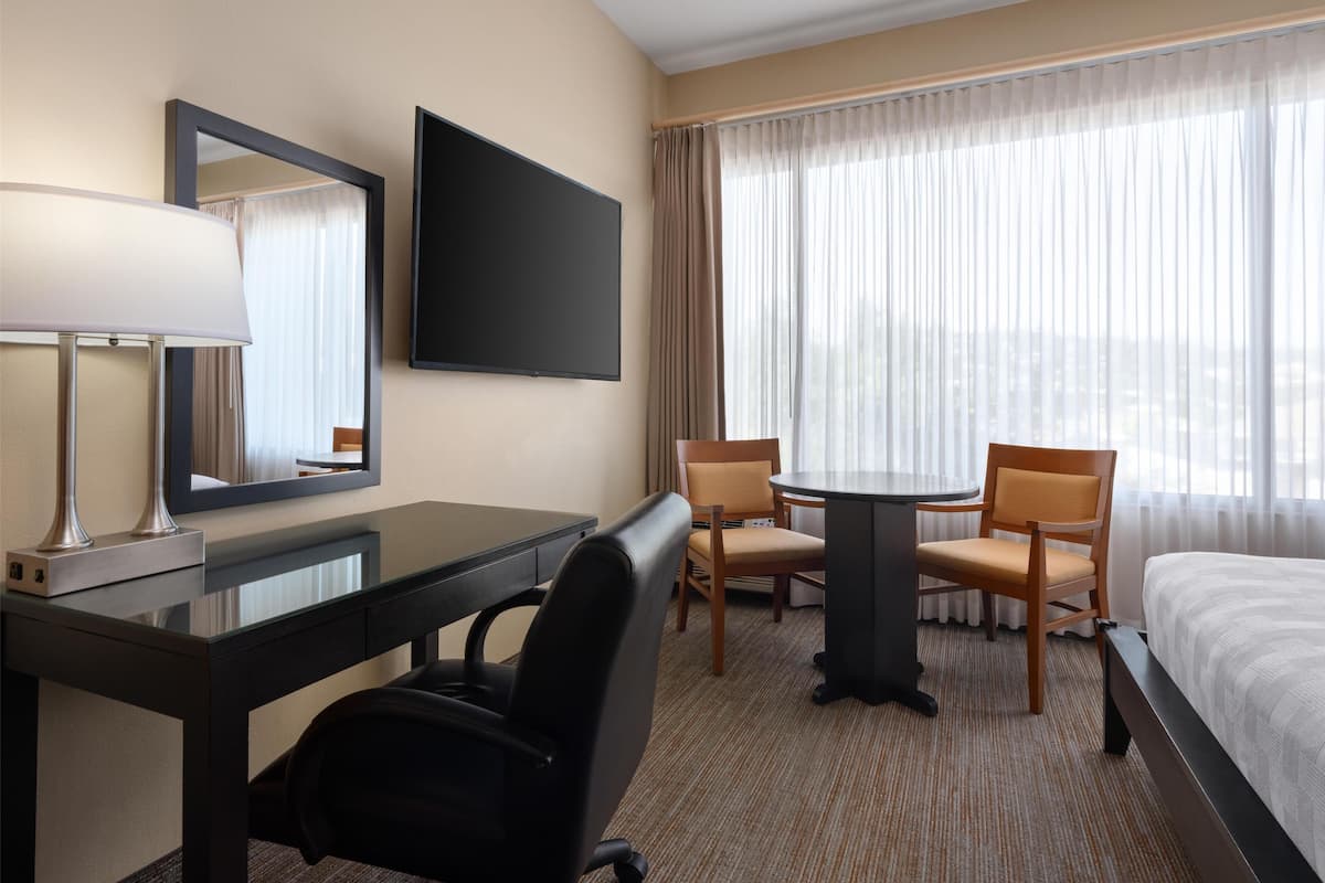 A hotel room with a bed, a desk with a chair, a TV, and a window with curtains.