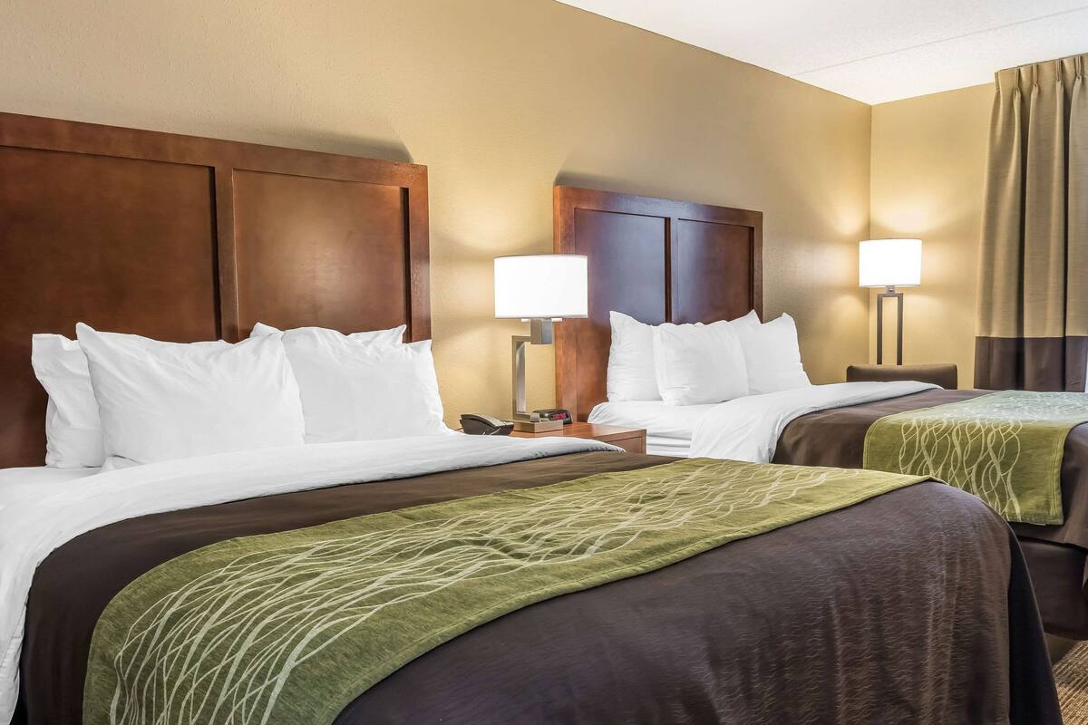 A hotel room with two beds, each with white linens and a green throw blanket. There are two bedside lamps and a nightstand with a phone.