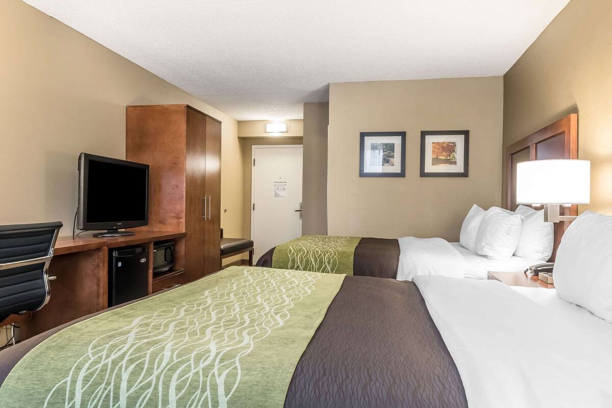 A hotel room with two beds, a desk with a television, a minibar, and a door leading to a hallway.