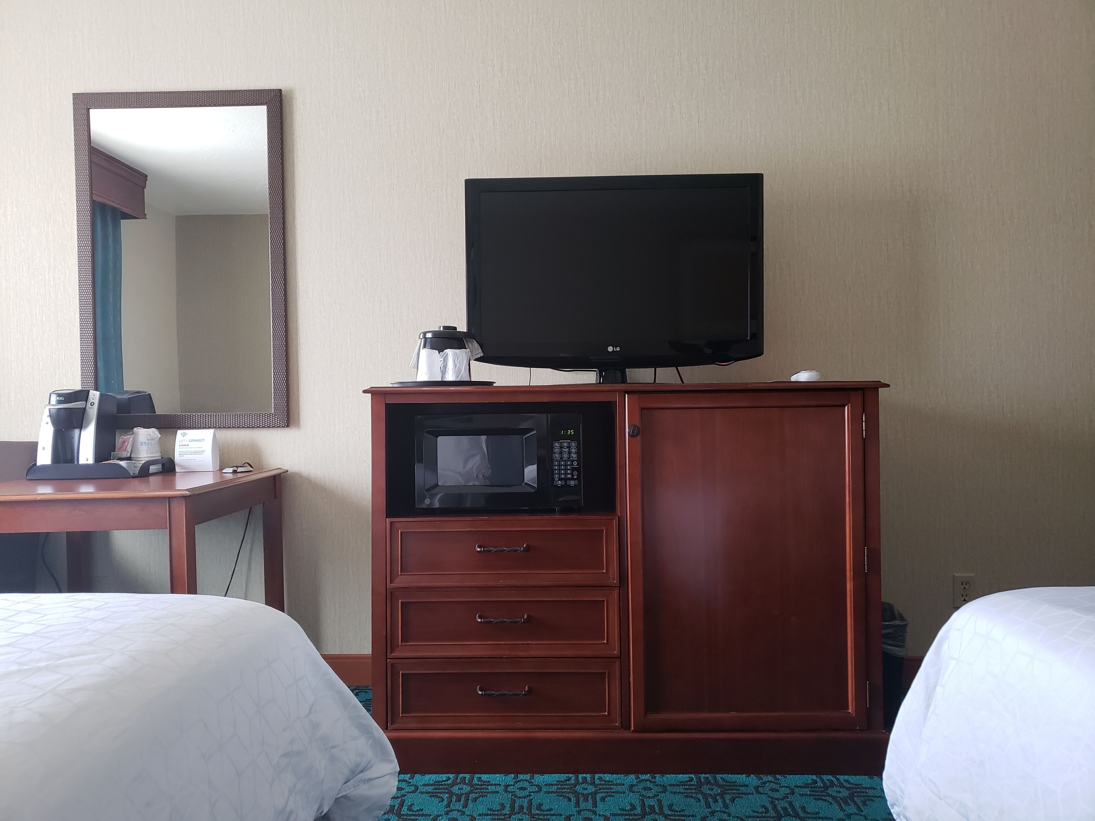 A hotel room with a wooden cabinet containing a microwave, a coffee maker, and a flat-screen TV. There is a mirror on the wall and two beds with white bedding.