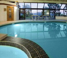 Una piscina interior con vistas despejadas al exterior gracias a amplios ventanales, una zona de solárium con baldosas y una puerta de madera con la etiqueta 'EXIT'.
