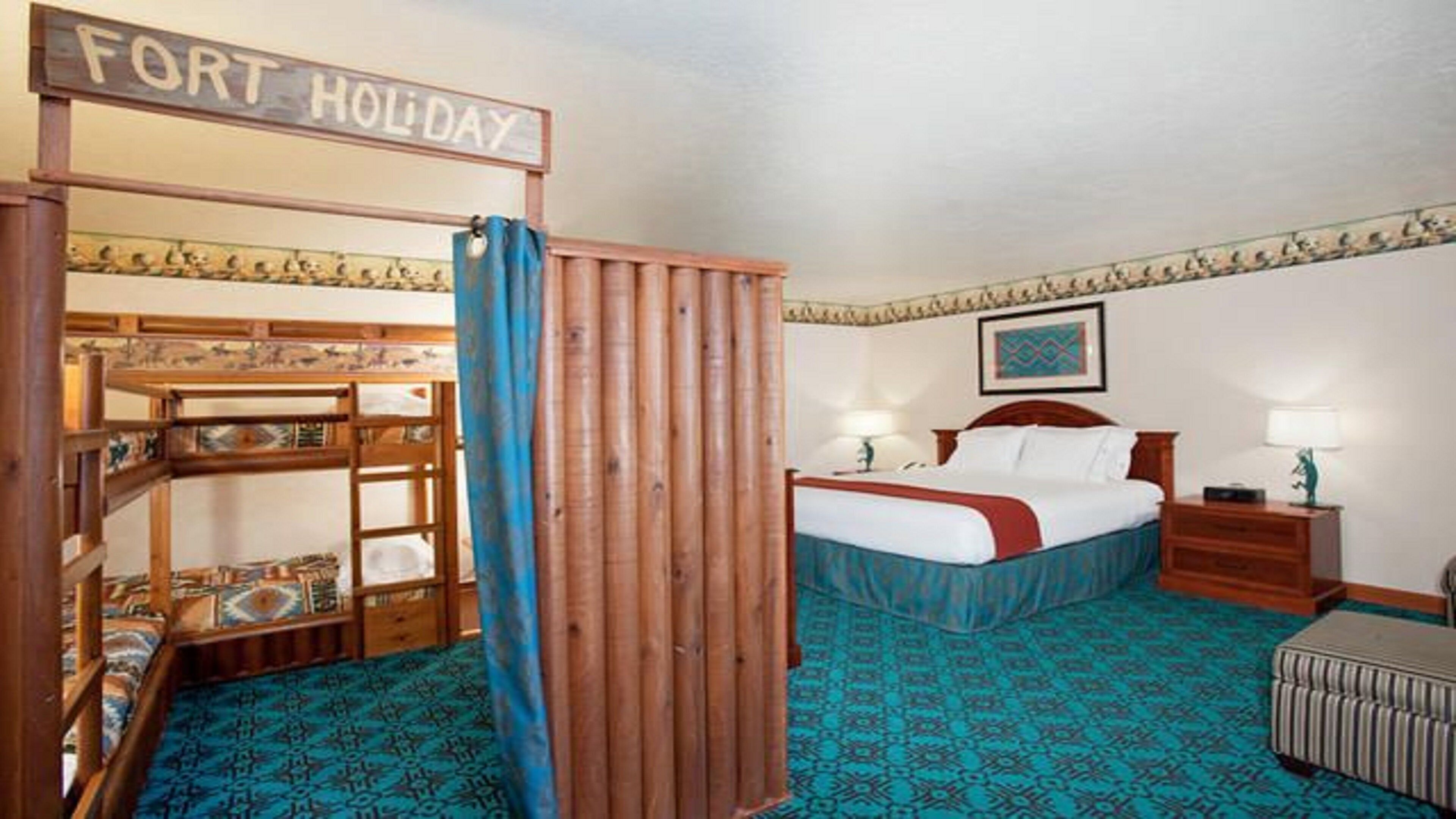 A hotel room with a bunk bed, a single bed, a nightstand, a lamp, and a sign that reads 'FORT HOLIDAY'.