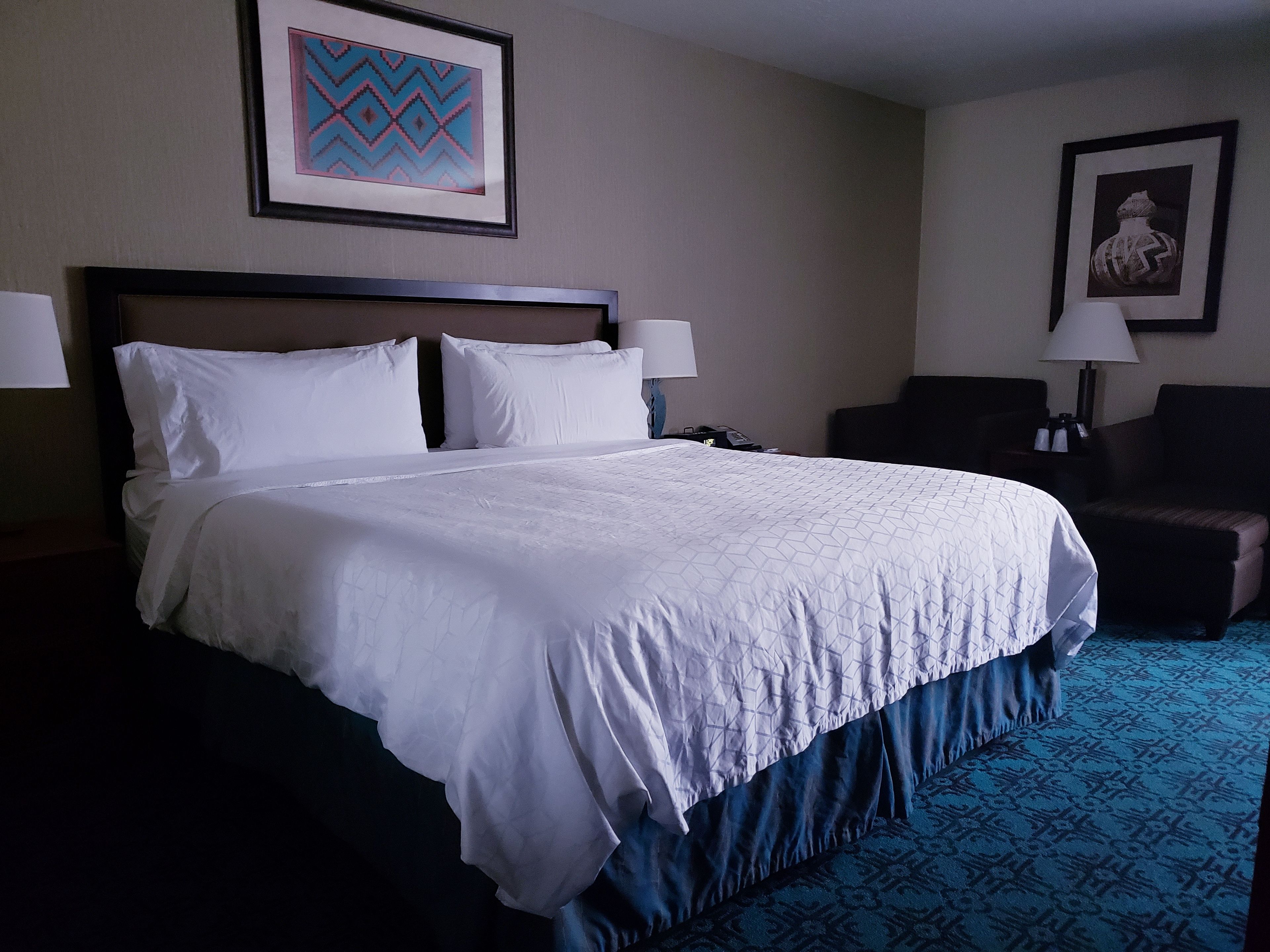 A hotel room with a large bed, two bedside lamps, a chair, and a framed picture on the wall.