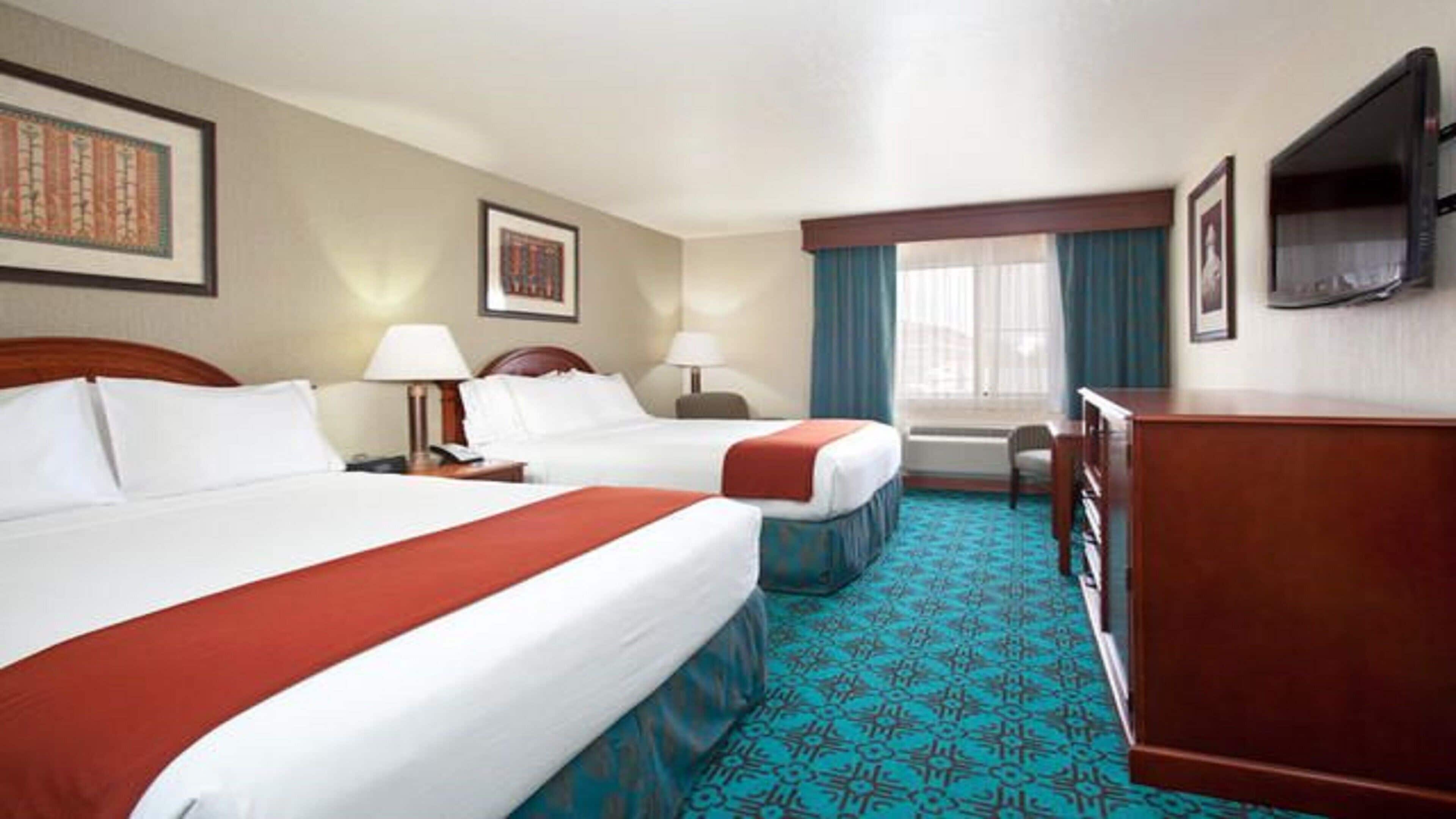 A hotel room with two beds, a desk, a TV, and a window with curtains.