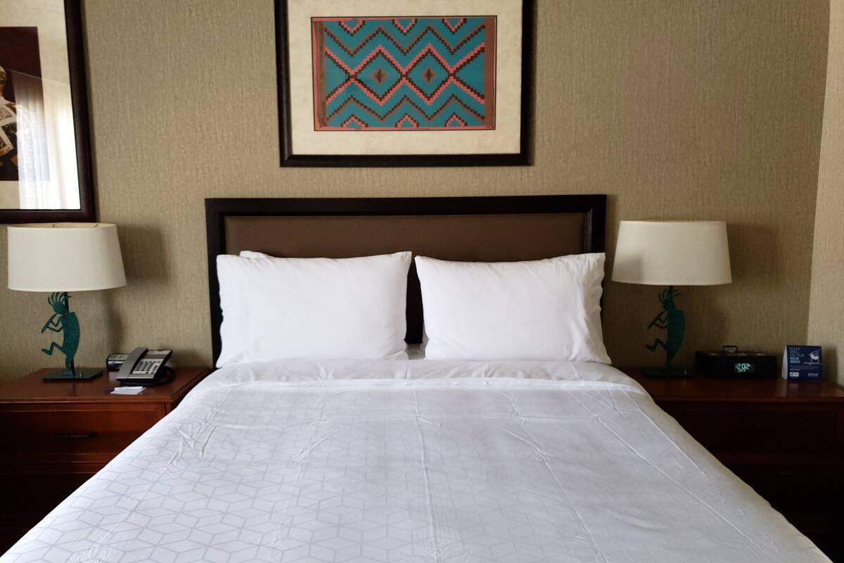 Une chambre d’hôtel avec un grand lit, deux tables de chevet avec des lampes, une œuvre d’art encadrée au-dessus du lit et une horloge sur l’une des tables.