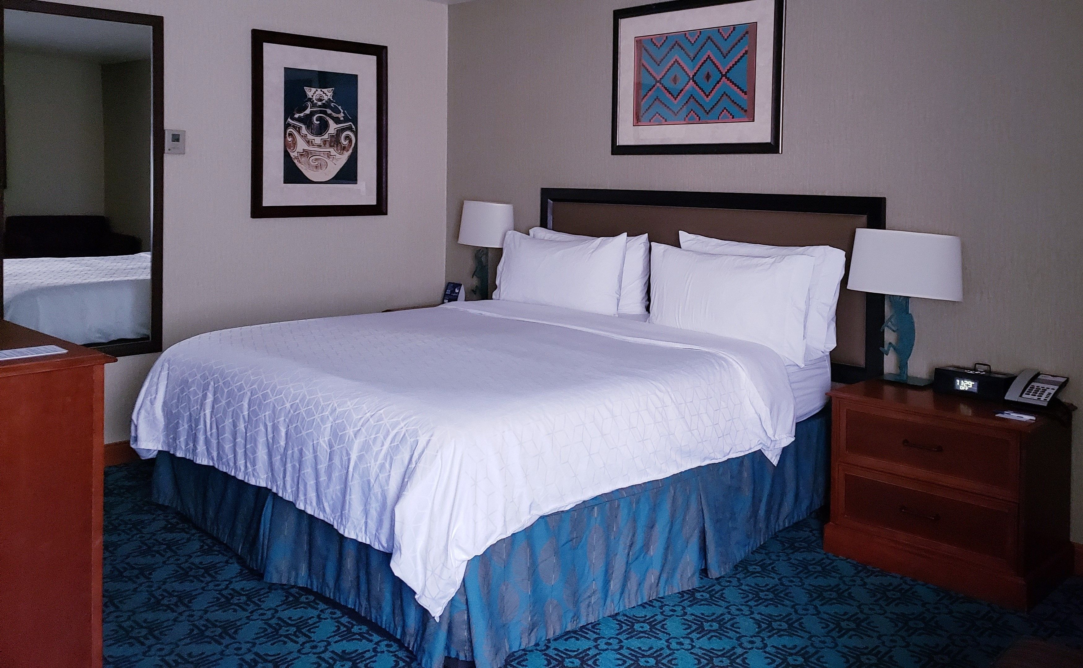 A neatly made bed with white linens, a wooden headboard, and a blue patterned bed skirt. There are two bedside tables with lamps and a telephone. The walls are adorned with framed artwork.