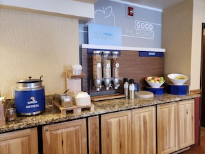 A breakfast bar with a 'Good Morning' sign, a 'Feelin' Fresh This Morning?' sign, and a 'Fruit' sign. There are containers of oatmeal, milk, and juice, as well as bowls of fruit. - Holiday Inn Express Mesa Verde-Cortez by IHG (Cortez)