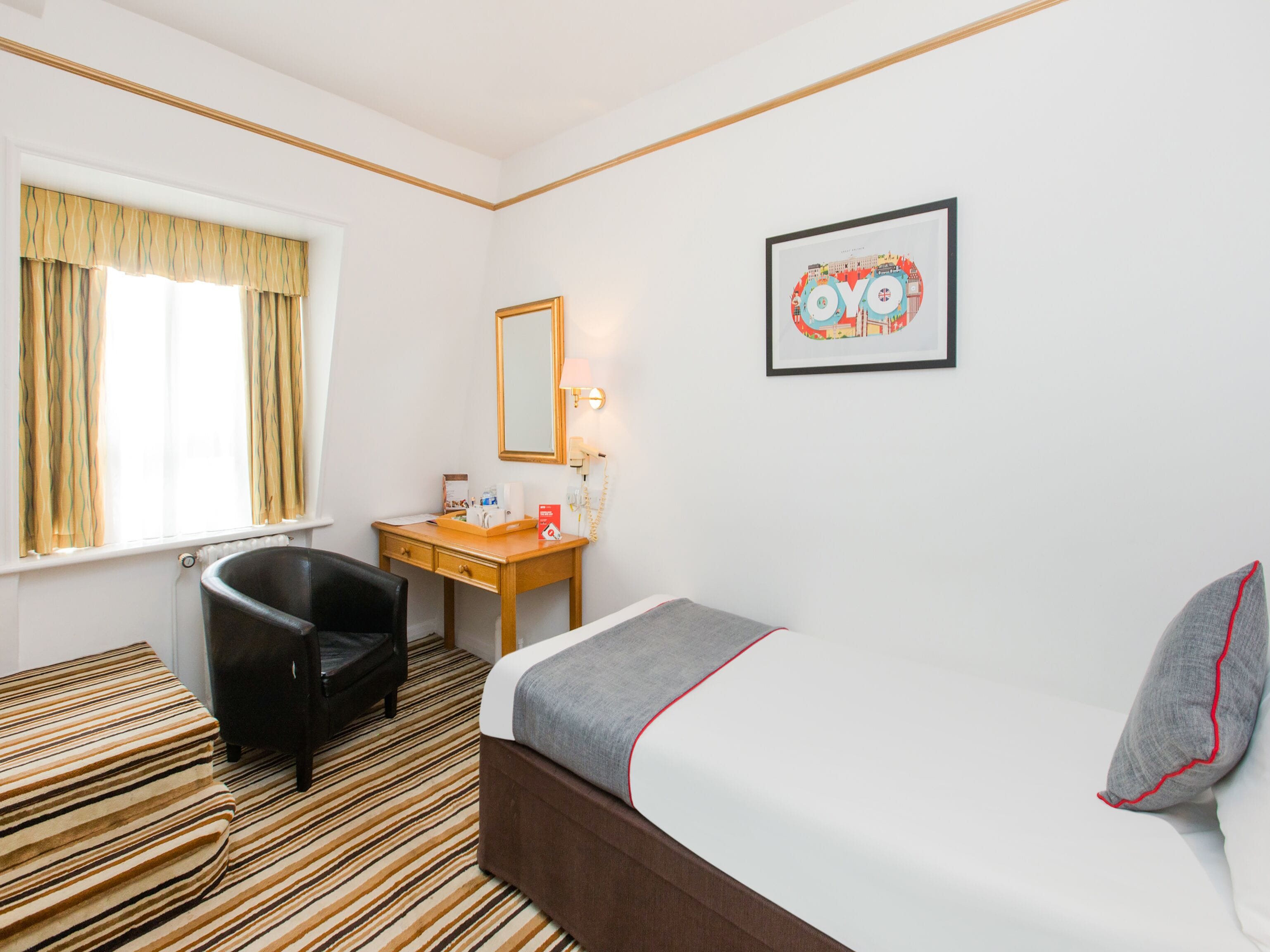 Une chambre d’hôtel comprenant un lit, un bureau, une chaise, une fenêtre avec des rideaux, un miroir fixé au mur et un tableau encadré.