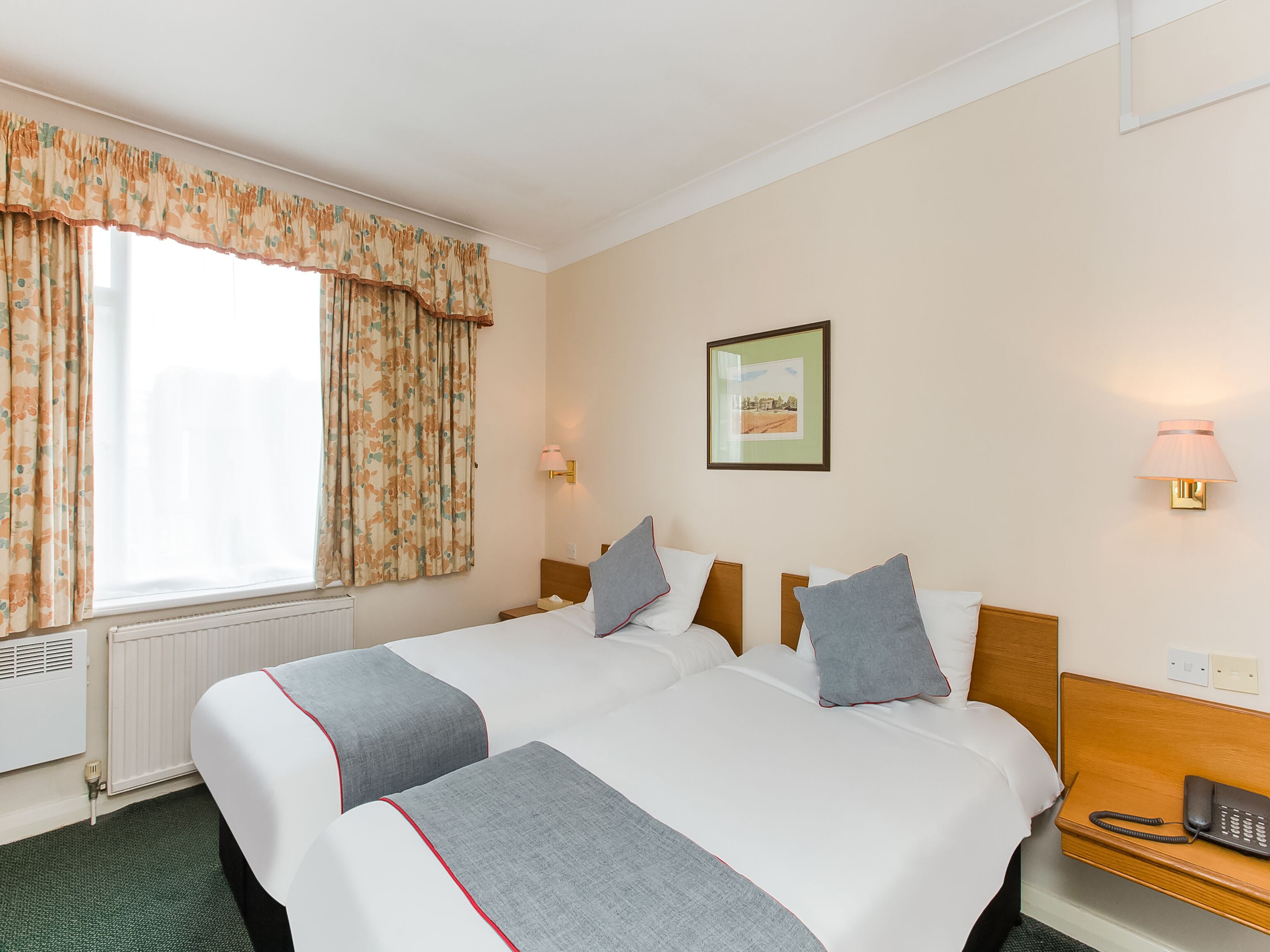 A hotel room with two beds, a window with curtains, a bedside lamp, a framed picture on the wall, and a phone on the nightstand.