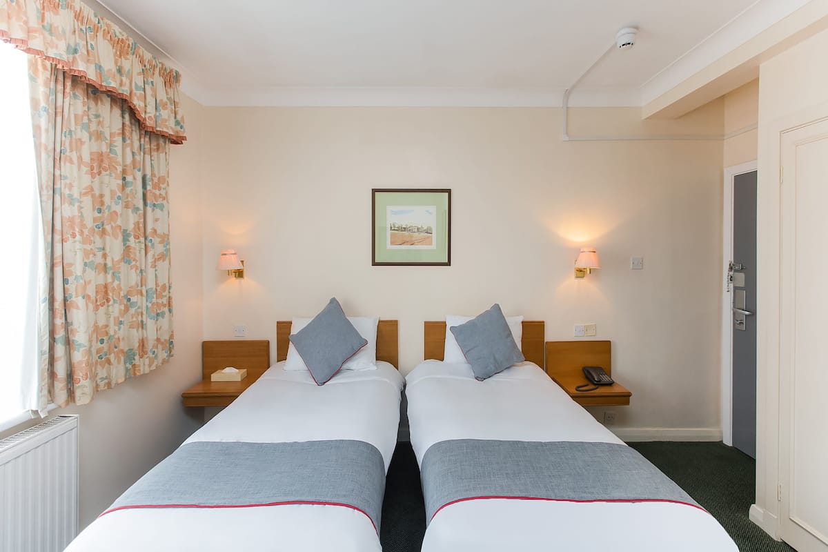 Habitación de hotel con dos camas individuales, cada una con una almohada y una mesita de noche. Se observa un cuadro en la pared, una ventana con cortinas y una puerta.