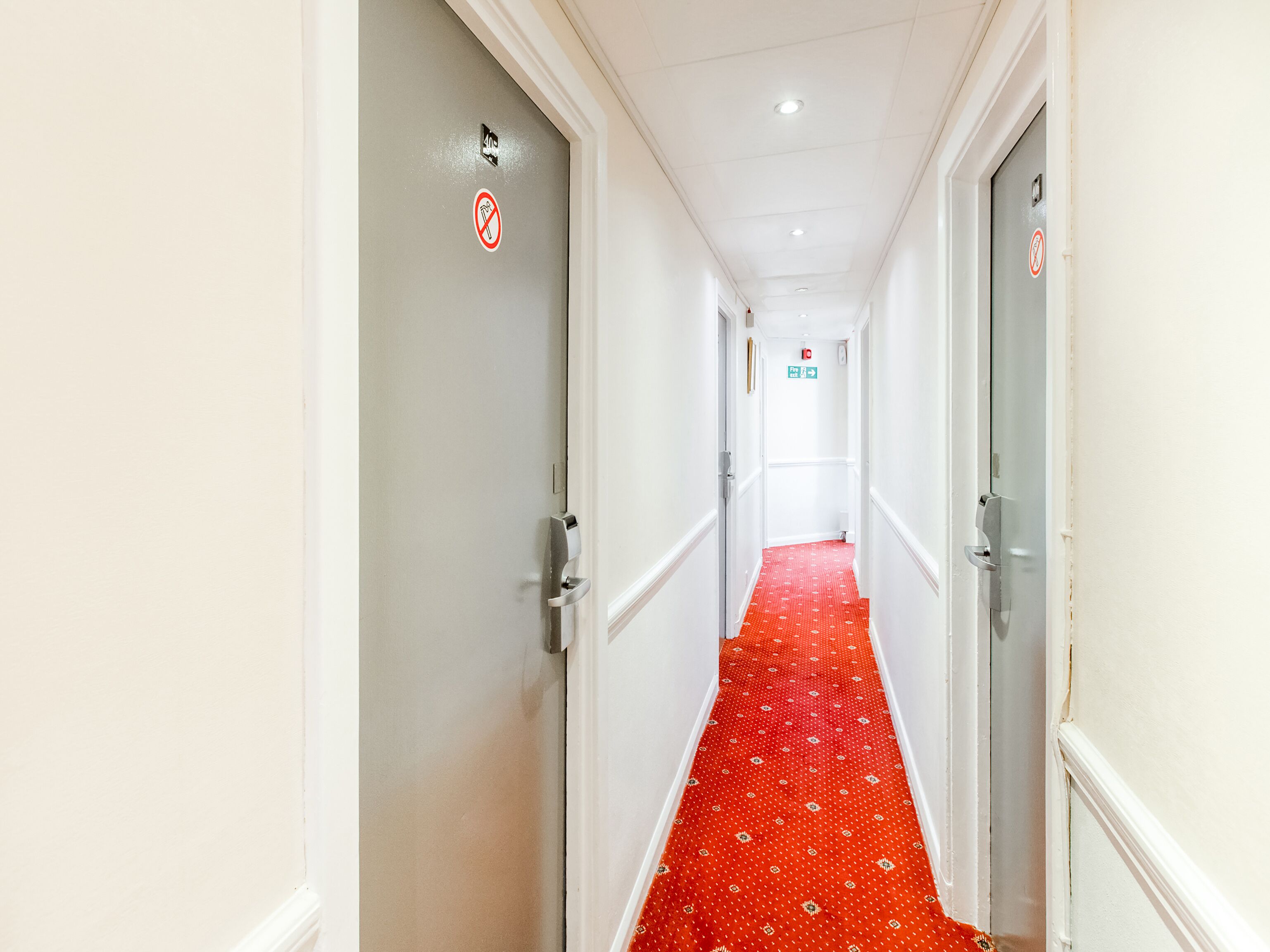 Un couloir avec un tapis rouge à motifs, des murs blancs et des portes ornées de panneaux « interdiction de fumer ».