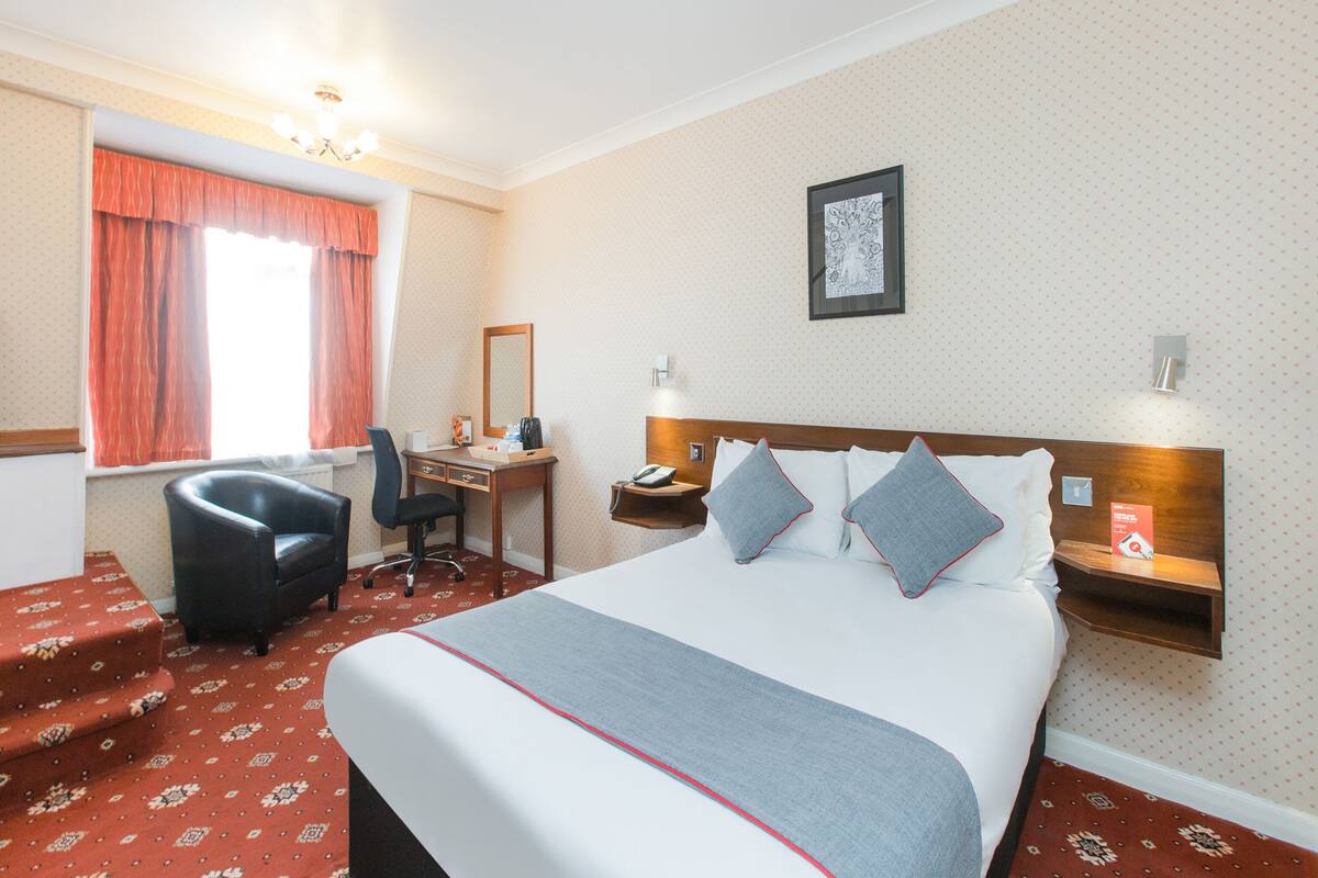 Habitación de hotel con una cama grande, un escritorio, una silla, una mesita de noche y un ventanal con cortinas.