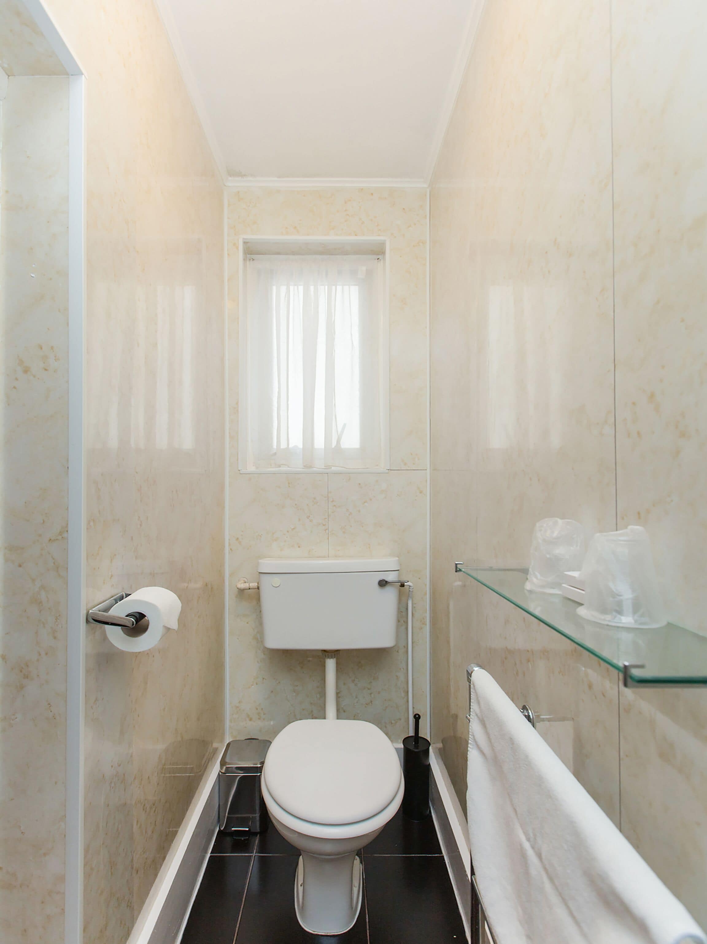 A small, modern bathroom with a toilet, a glass shelf, and a window with curtains.