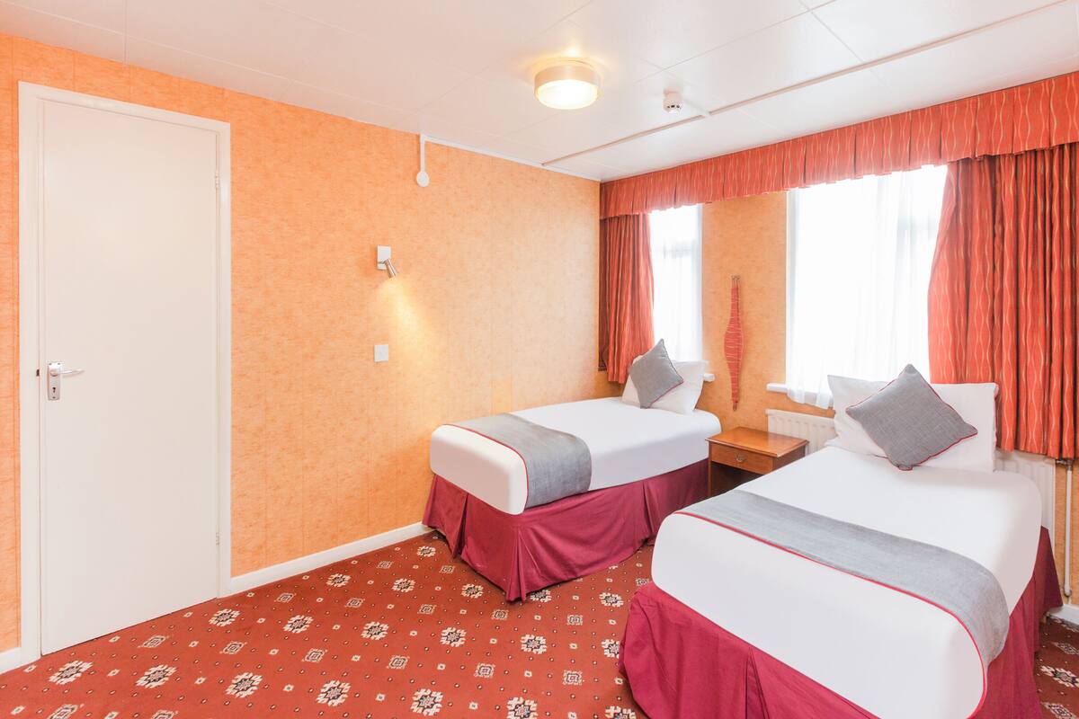 Habitación de hotel con dos camas, una puerta blanca, alfombra roja estampada y una ventana con cortinas rojas.