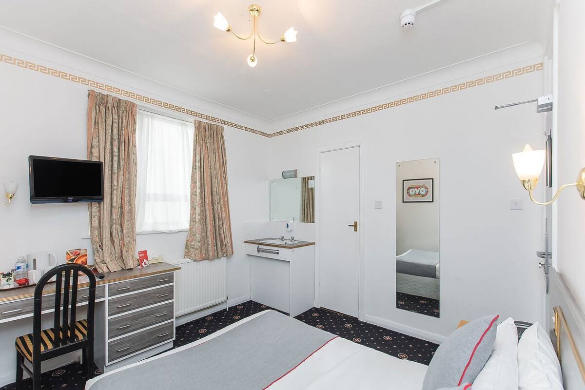 Habitación de hotel con una cama, un escritorio con silla, televisión, espejo y ventana con cortinas.