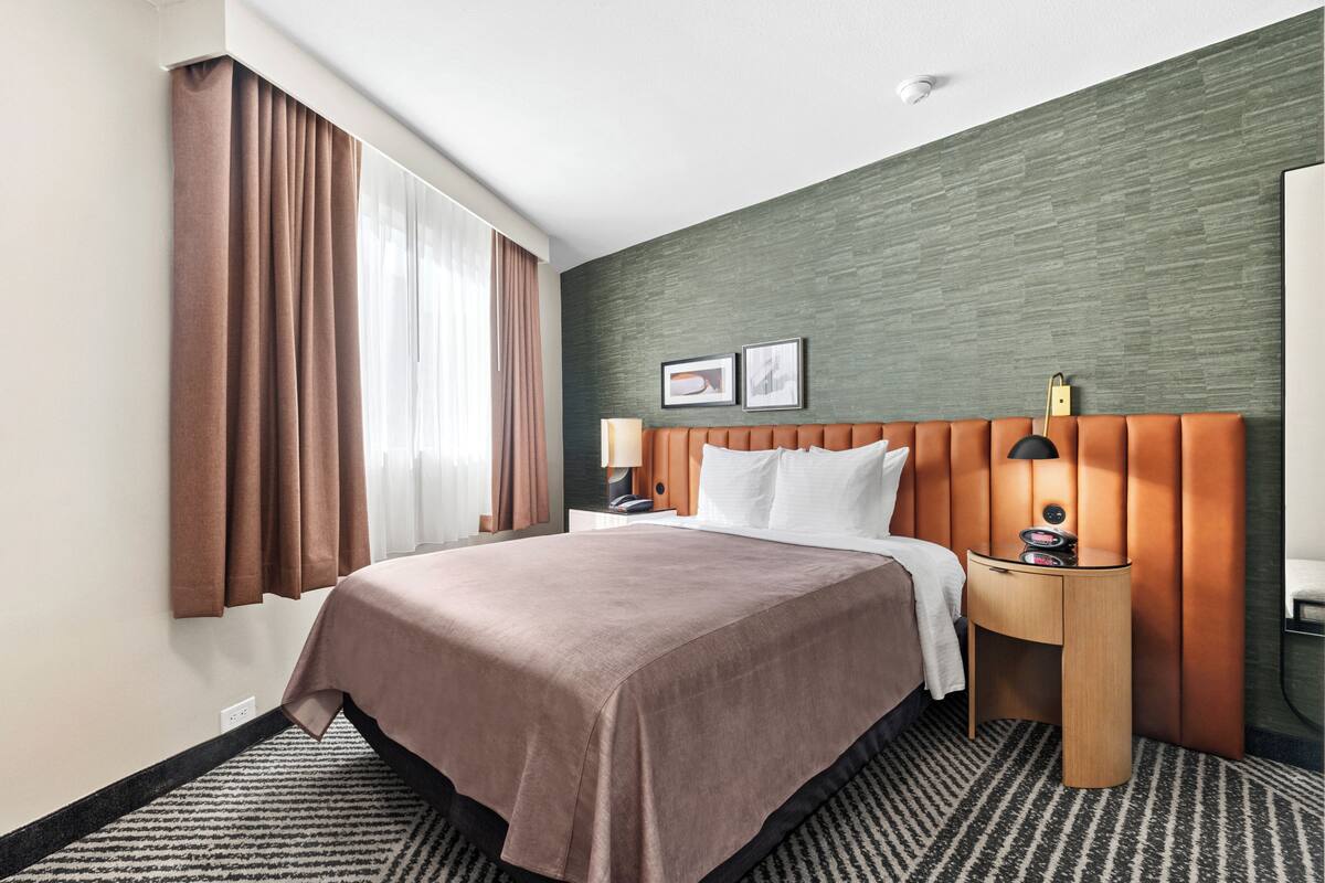 Un cuarto de hotel con una cama grande, un cabecero de madera, una mesita de noche con una lámpara, y vistas al exterior por la ventana.