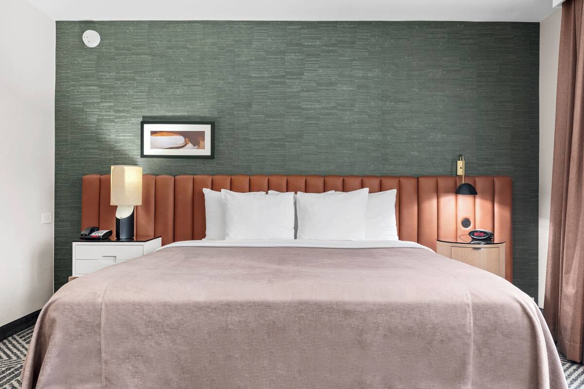 Een hotelkamer met een groot bed, een nachtkastje met lamp, een wandklok en een lijst met een afbeelding boven het bed.