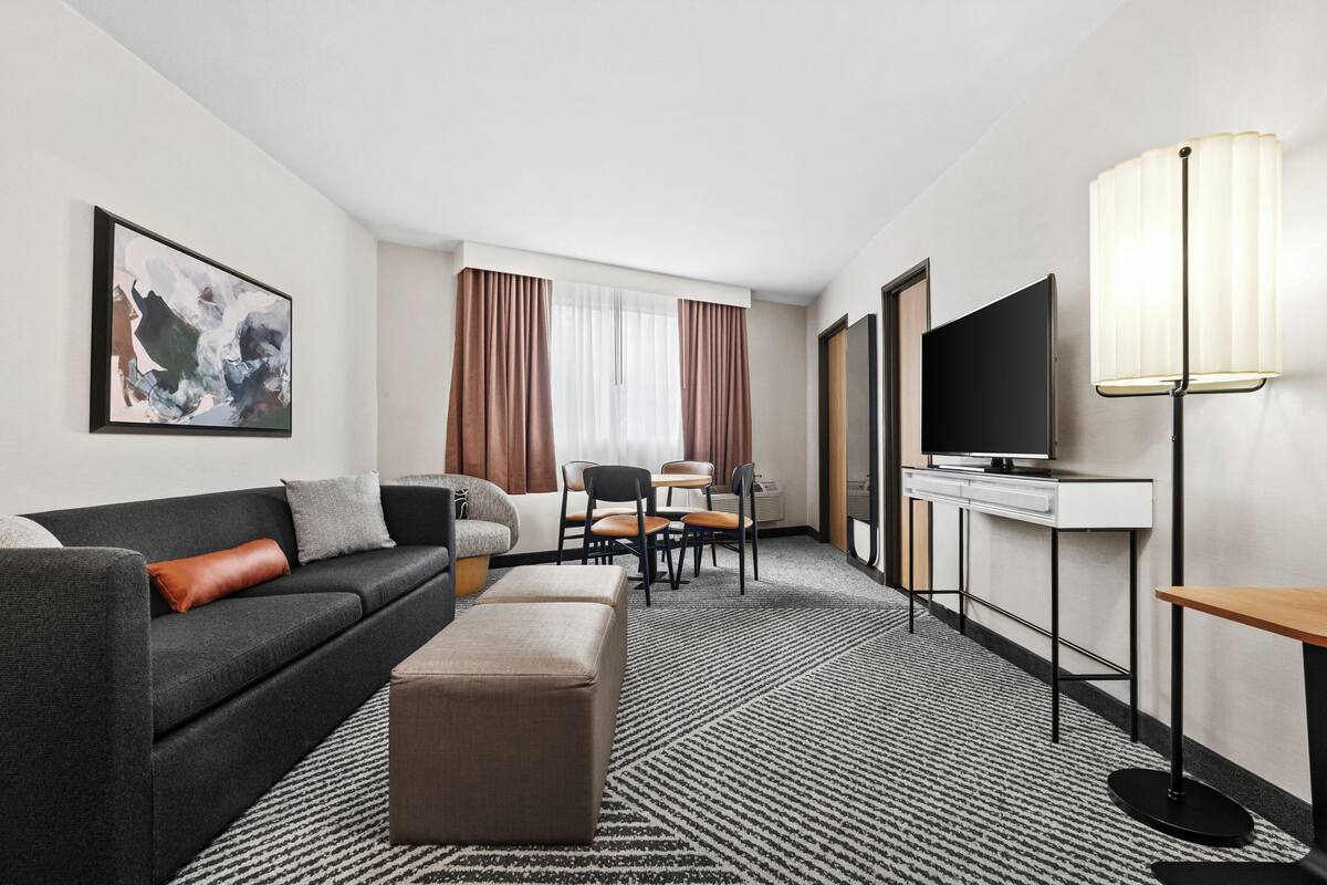 Una habitación de hotel con sofá, reposapiés, televisión, lámpara y un área de comedor con sillas y mesa.