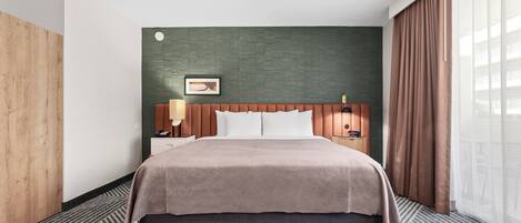Habitación de hotel con una cama grande, un armario de madera, una mesita de noche con lámpara y vistas a la ciudad a través de amplios ventanales.