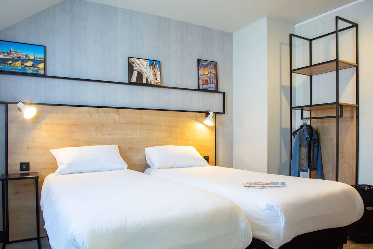 Une chambre dâhĂŽtel avec deux lits, une tĂȘte de lit en bois, une Ă©tagĂšre avec des tableaux, un porte-manteau et une table de chevet avec une lampe.