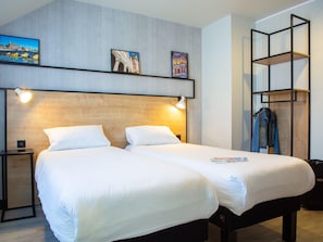Une chambre d’hôtel avec deux lits, une tête de lit en bois, une étagère avec des tableaux, un porte-manteau et une table de chevet avec une lampe.