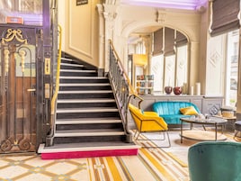 Un escalier menant à un hall d’entrée où se trouvent un canapé bleu, un fauteuil jaune et une petite table avec une lampe.