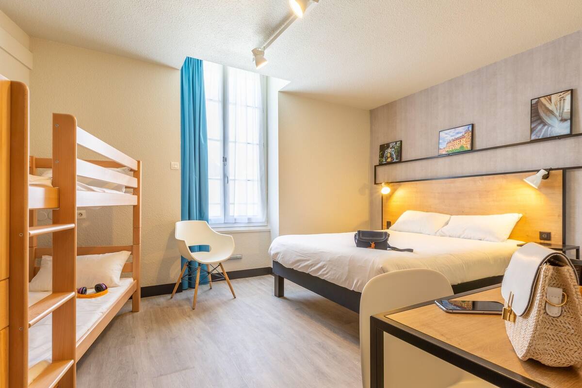 Une chambre d’hôtel avec un lit superposé, un bureau avec une chaise, une table de chevet avec une lampe, une fenêtre avec des rideaux et un lit avec un oreiller et un sac.