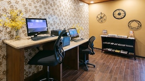 A reception desk with two monitors, a printer, and a computer. There are two chairs, a vase with yellow flowers, and a bookshelf with magazines and a box labeled "The Bee".