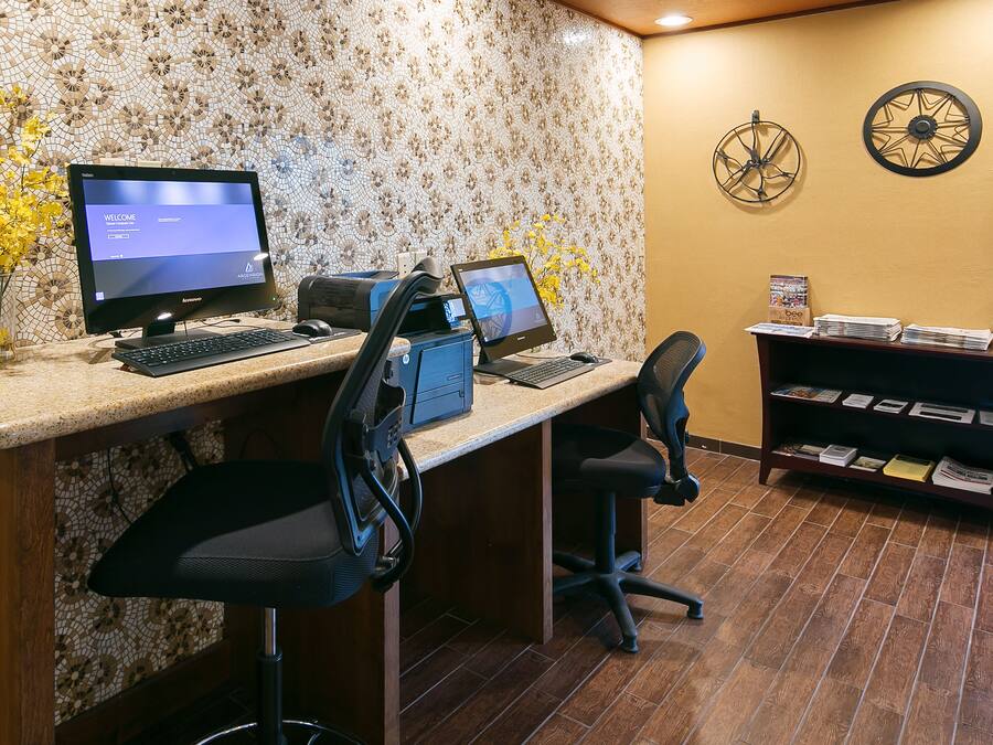 A reception desk with two monitors, a printer, and a computer. There are two chairs, a vase with yellow flowers, and a bookshelf with magazines and a box labeled "The Bee".