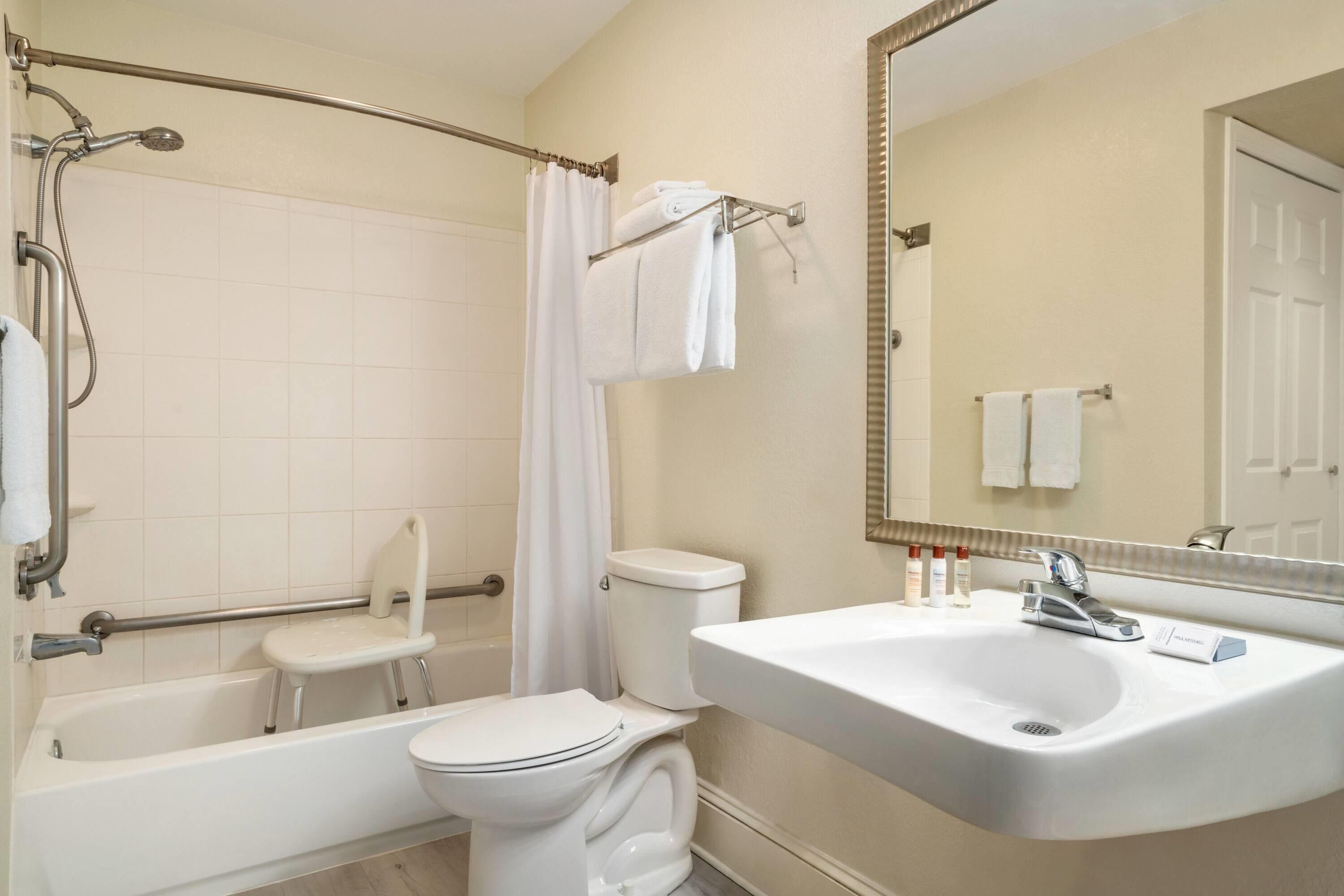 Un baño con bañera, inodoro y lavabo, equipado con una silla de ducha y barras de apoyo.