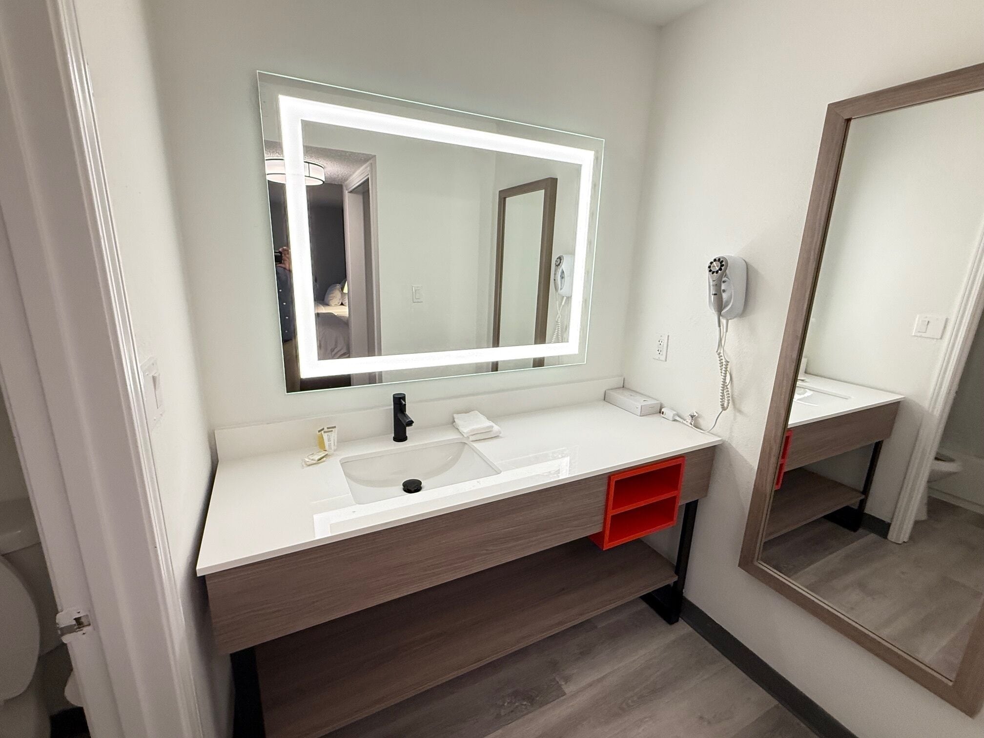 Un baño moderno con un tocador blanco, un espejo grande con iluminación integrada, un secador de pelo y un cajón de almacenamiento rojo.
