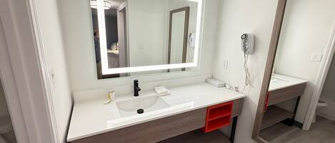 Un baño moderno con un tocador blanco, un espejo grande con iluminación integrada, un secador de pelo y un cajón de almacenamiento rojo.
