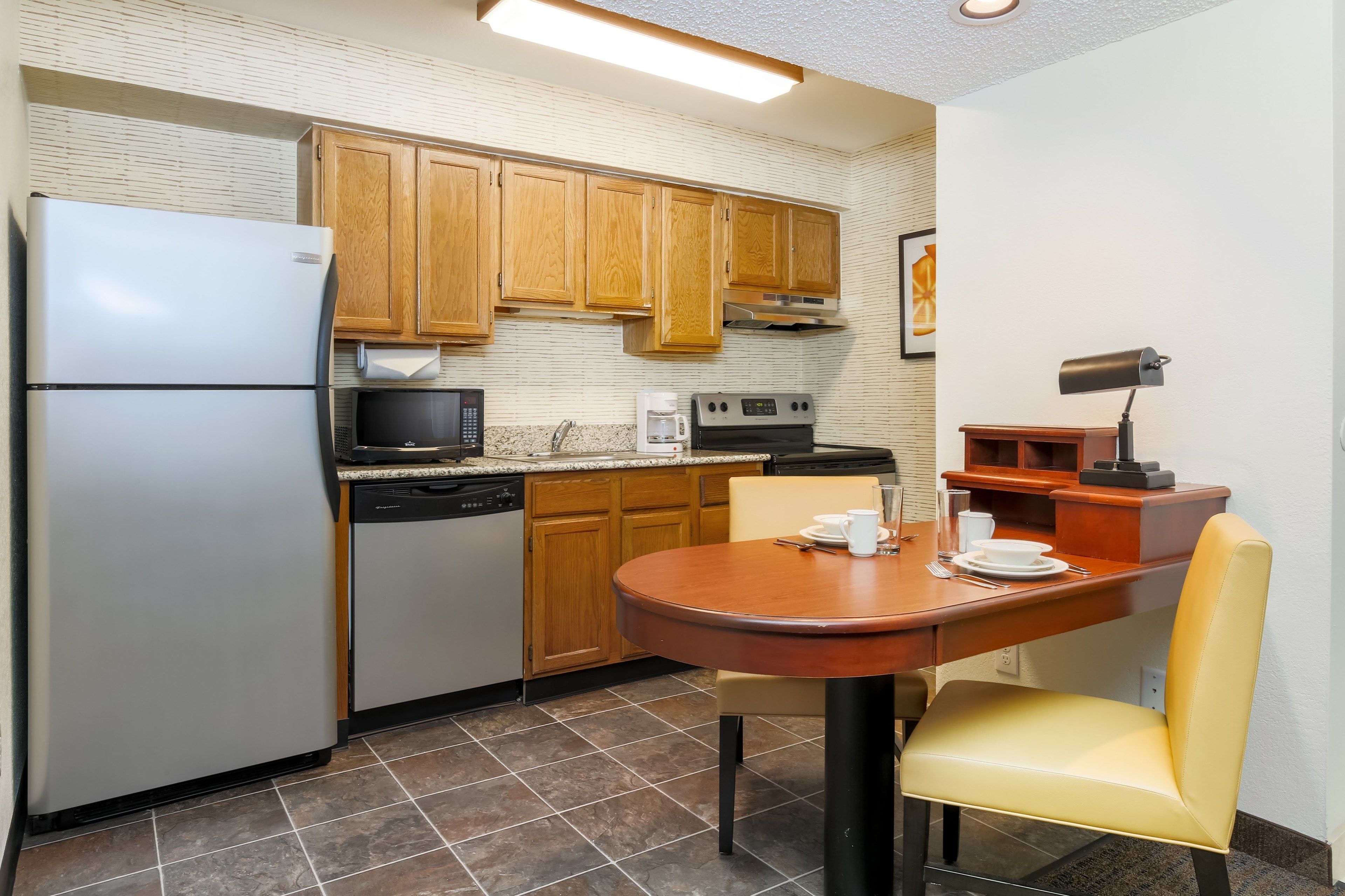 A compact kitchen with wooden cabinets, a refrigerator, microwave, coffee maker, and a dining table with chairs.