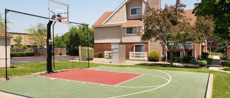 An outdoor basketball court with a red and green surface, surrounded by a fence and trees.