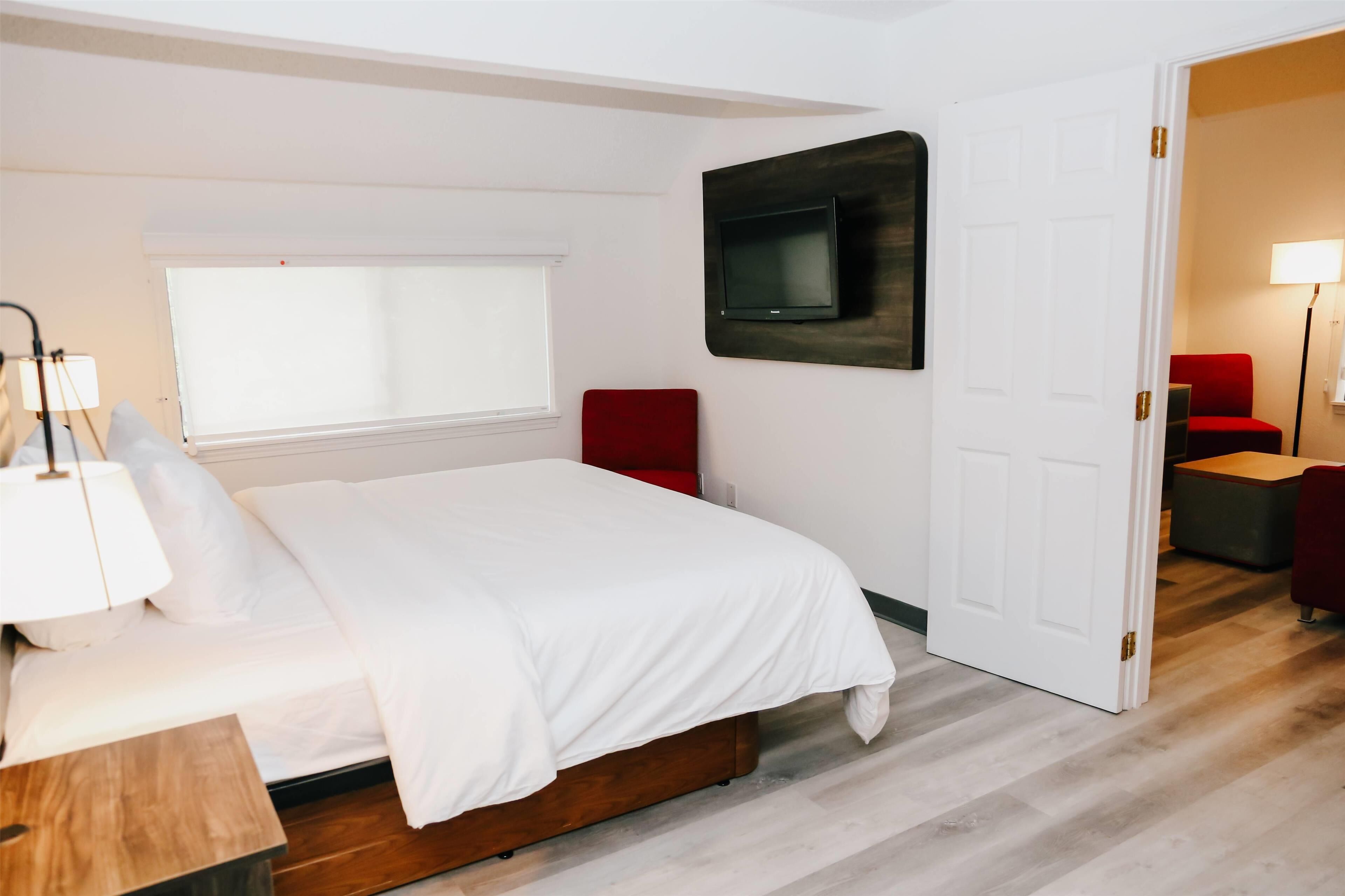 A bedroom with a bed, a TV mounted on the wall, a window with blinds, a red chair, and a door leading to another room.