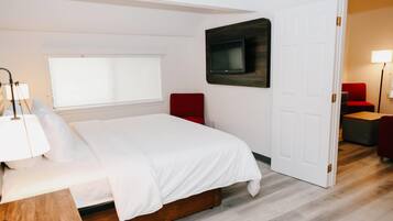 A bedroom with a bed, a TV mounted on the wall, a window with blinds, a red chair, and a door leading to another room.