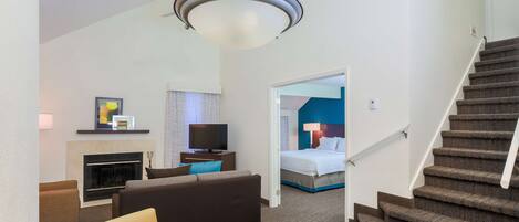 Una habitación de hotel con sala de estar, chimenea, televisor, cama y escalera.