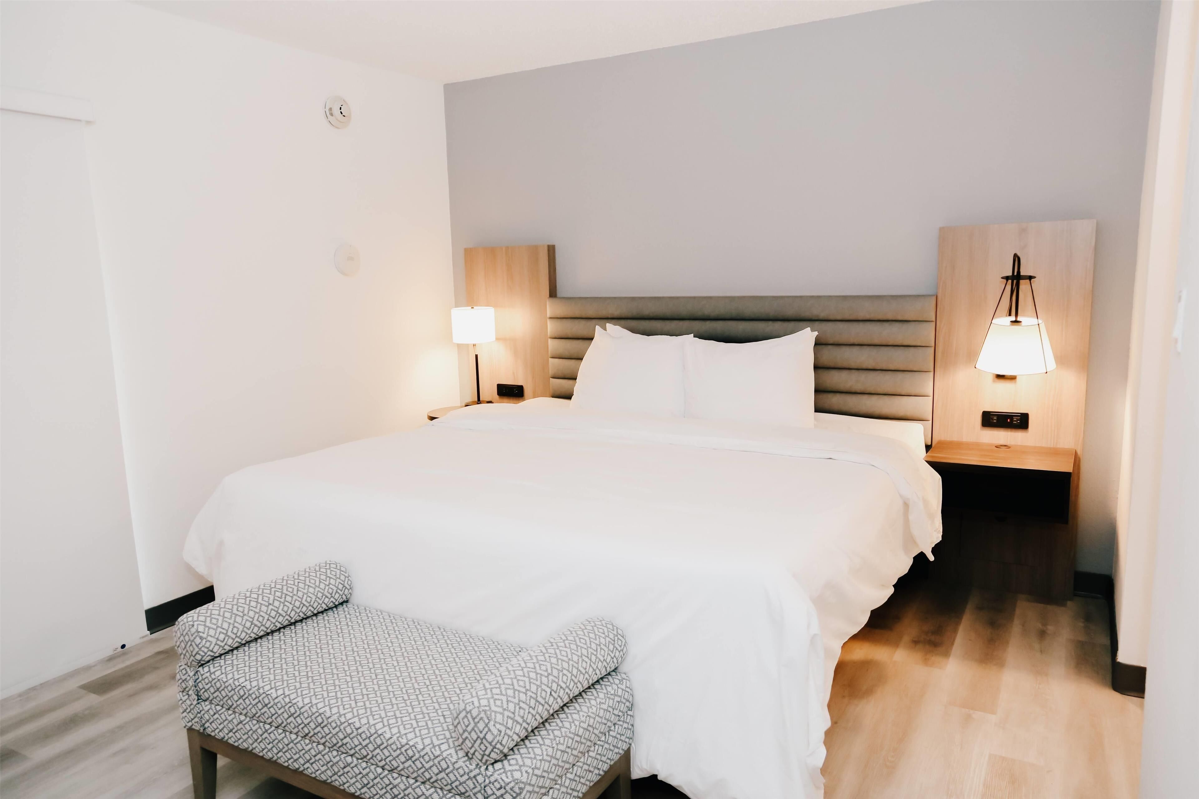 Ein ordentlich eingerichtetes Hotelzimmer mit einem großen Bett, zwei Nachttischen mit Lampen, einer Bank und einem Holz-Kopfteil.