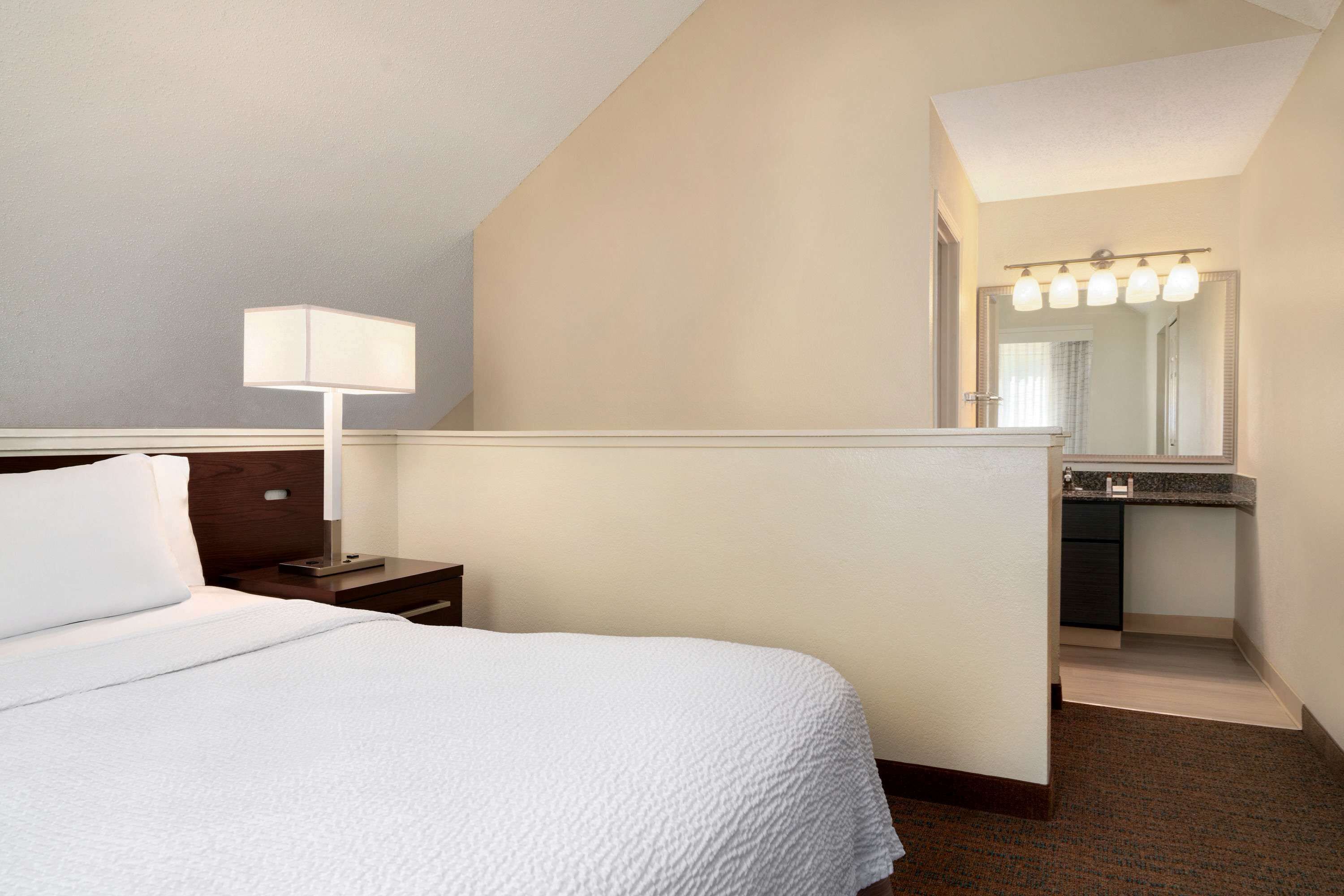 A hotel room with a bed, a nightstand, a lamp, a bathroom with a mirror and lights, and a carpeted floor.