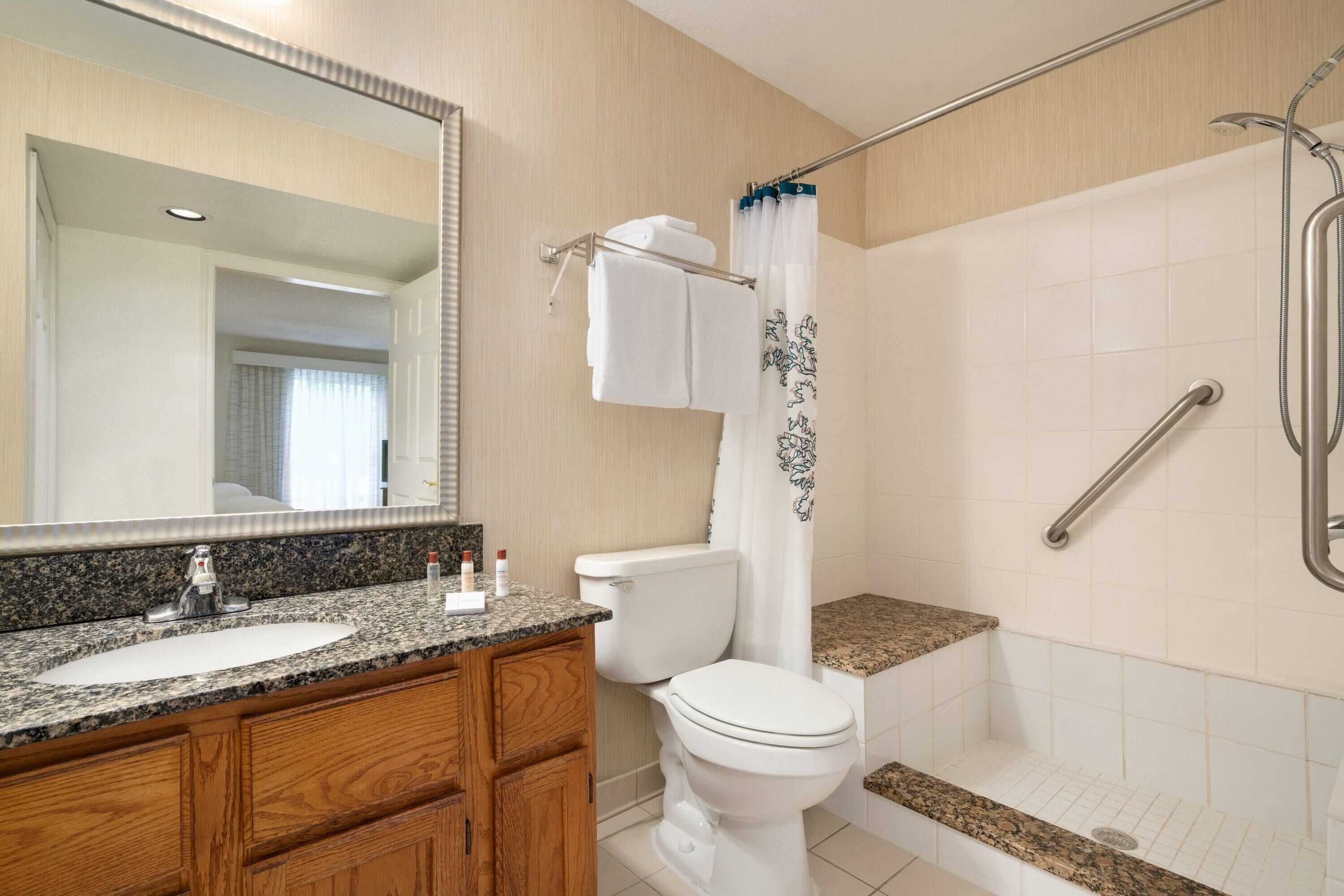 A bathroom with a granite countertop sink, wooden vanity, a white toilet, a shower with a grab bar, and a large mirror.