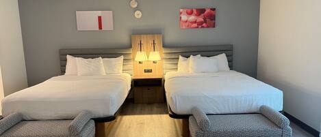 A hotel room with two beds, a wooden headboard, bedside tables with lamps, a bench, and a wall-mounted artwork.
