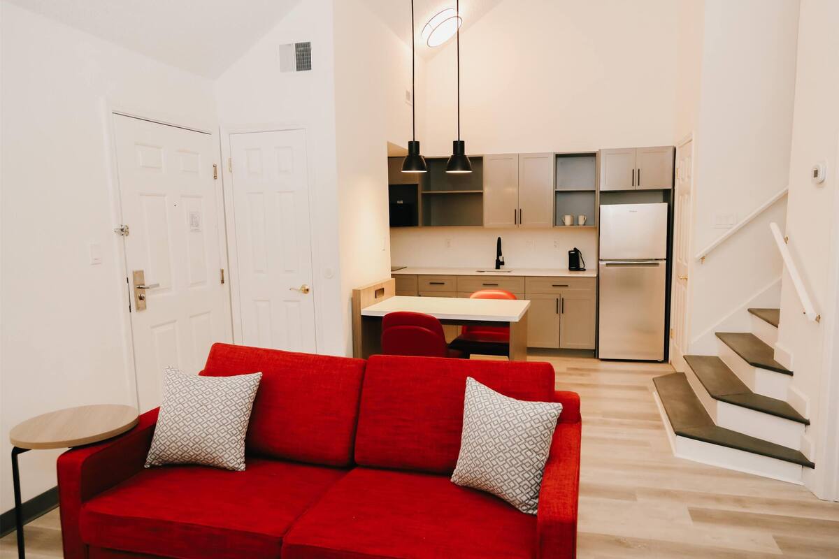 Un espacio habitable compacto con un sofá rojo, una mesa de comedor, una zona de cocina y una escalera.