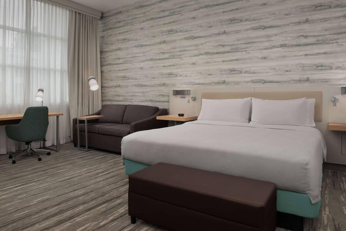 A hotel room with a large bed, a desk, two chairs, a small table, and a window with curtains.