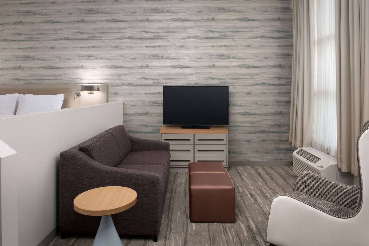 A modern hotel room with a grey sofa, a small round wooden table, a brown ottoman, a flat-screen TV on a wooden stand, and a wicker chair.