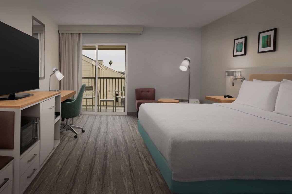 A hotel room with a bed, desk, chair, and a balcony with a view.